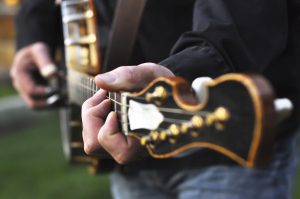 A closeup on a man's left hand playing a Banjo. The Banjo is out of focus except for the neck where his hands are playing a chord. The Banjo hangs from a leather strap around his body. His head is not shown.